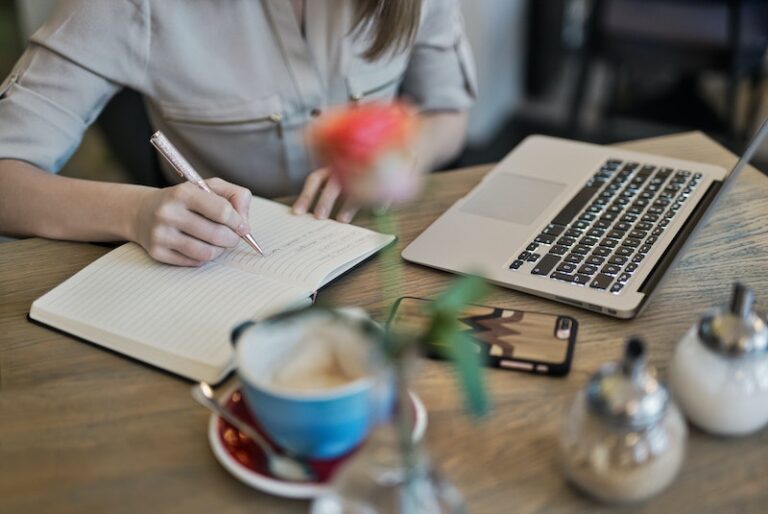 A person working at a desk - writing in a note book with a laptop open and cup of coffee