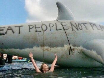 Amelia Crews swimming in the ocean in front of a large inflatable shark with the words Eat People Not Plastic