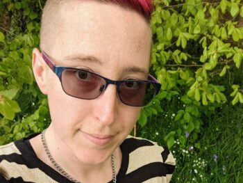 Alexis with pink hair, sunglasses and a striped top smiling at the camera