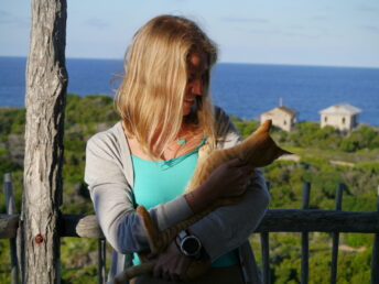 Melissa holding a ginger cat and standing on a wooden balcony with the blue ocena behind her