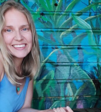Melissa wearing a blue top and smiling at the camera with a blue wall and painted mural of jungle plants in the background