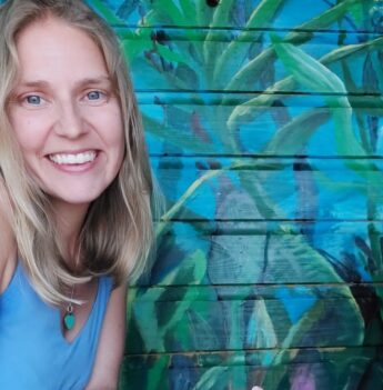Melissa wearing a blue top and smiling at the camera with a blue wall and painted mural of jungle plants in the background