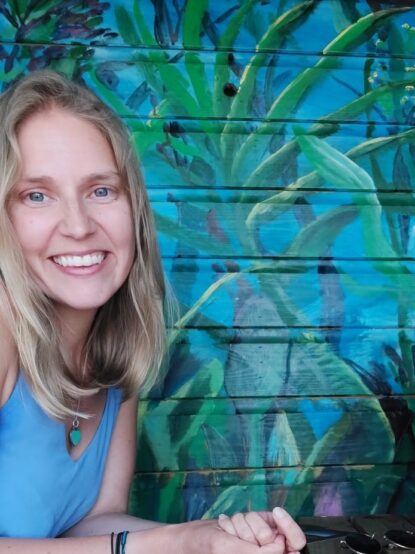 Melissa wearing a blue top and smiling at the camera with a blue wall and painted mural of jungle plants in the background {{brizy_dc_image_alt imageSrc=