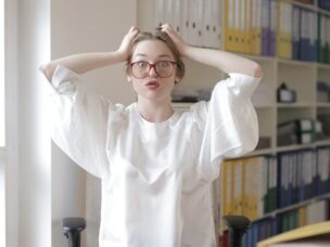 A woman standing in an office wearing a white top and red glasses stares at the camera and gestures as if to tear her hair out in frustration